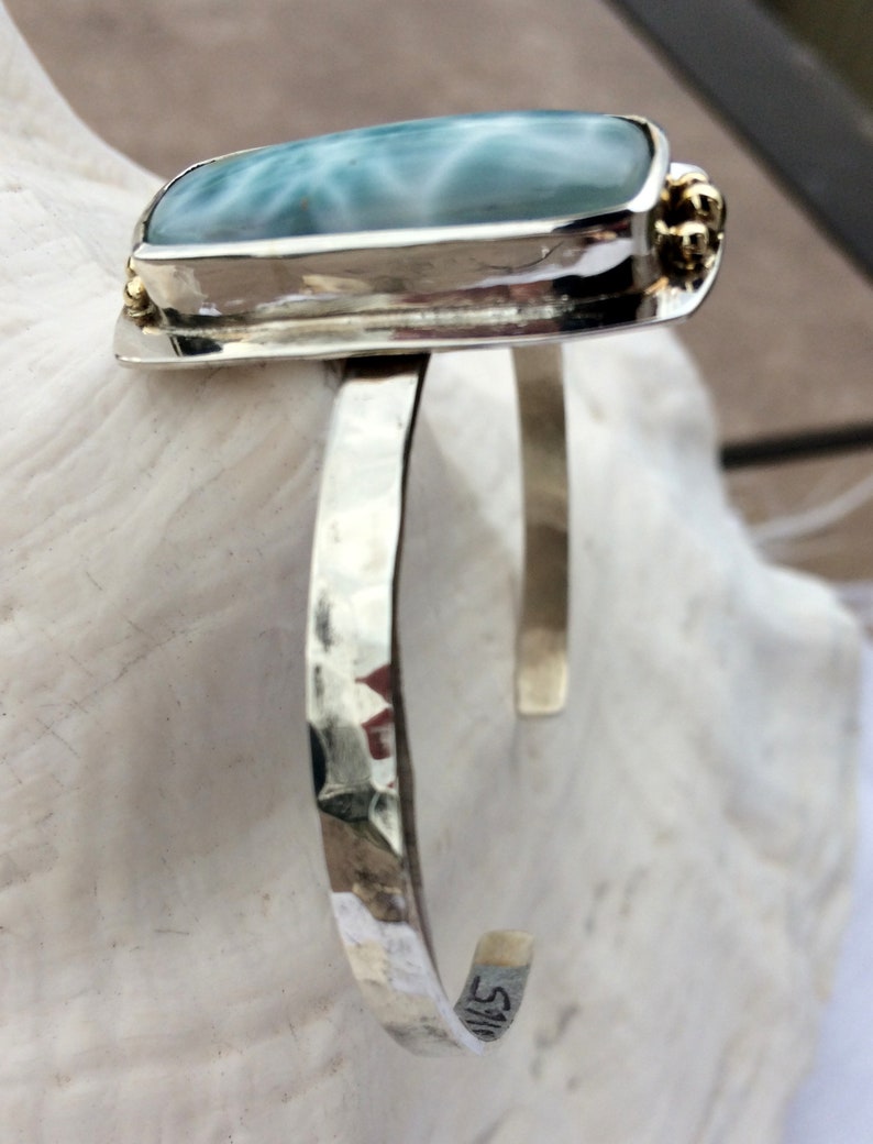 May include: A silver cuff bracelet featuring a light blue and white gemstone set in a silver bezel. The bracelet has a hammered finish and is accented with small gold beads.