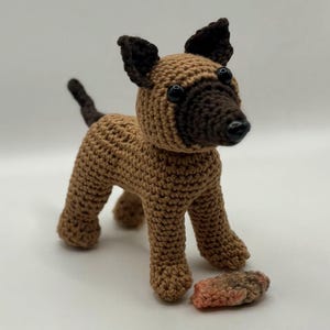 Könnte beinhalten: Gehäkelte Hundefigur in Braun- und Dunkelbrauntönen, mit schwarzen Augen und einem kleinen Schwanz. Der Hund steht neben einem kleinen, mehrfarbigen gehäkelten Objekt. Die Ohren und die Schnauze des Hundes sind dunkler braun.