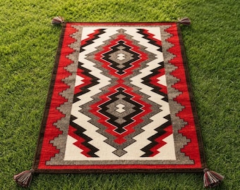 May include: A rectangular woven rug featuring a geometric pattern in red, black, white, and gray. The design consists of repeating diamond and zigzag shapes centered on the rug, with a dark border and small tassels attached to each of the four corners.