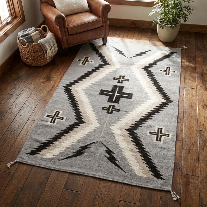 Puede incluir: Una alfombra rectangular gris con un patr&oacute;n geom&eacute;trico. El dise&ntilde;o presenta zigzags, cruces y formas de rel&aacute;mpagos en negro, blanco y beige. La alfombra tiene borlas en los extremos y est&aacute; colocada sobre un piso de madera noble.