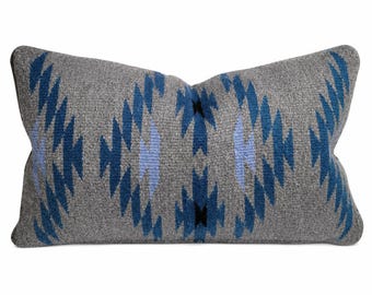 Puede incluir: Un cojín rectangular con una base de color gris y un patrón geométrico tejido en tonos azul oscuro, azul claro y negro. El diseño consiste en formas repetitivas de diamantes y chevrones en toda la superficie frontal del cojín.