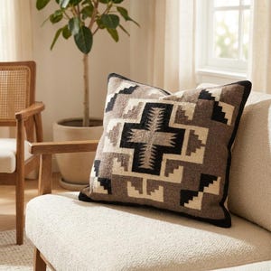 May include: Square throw pillow with a geometric pattern in shades of brown, black, and cream. The pillow has a black border and is placed on a cream-colored sofa. The pillow is a decorative home accent.