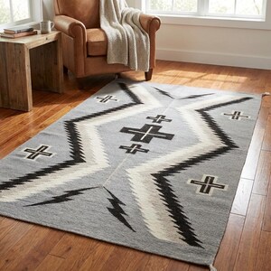 Puede incluir: Una alfombra rectangular gris con un patr&oacute;n geom&eacute;trico. La alfombra presenta l&iacute;neas en zigzag negras, blancas y grises y formas de cruz. La alfombra est&aacute; hecha de un material tejido y tiene flecos en los extremos.