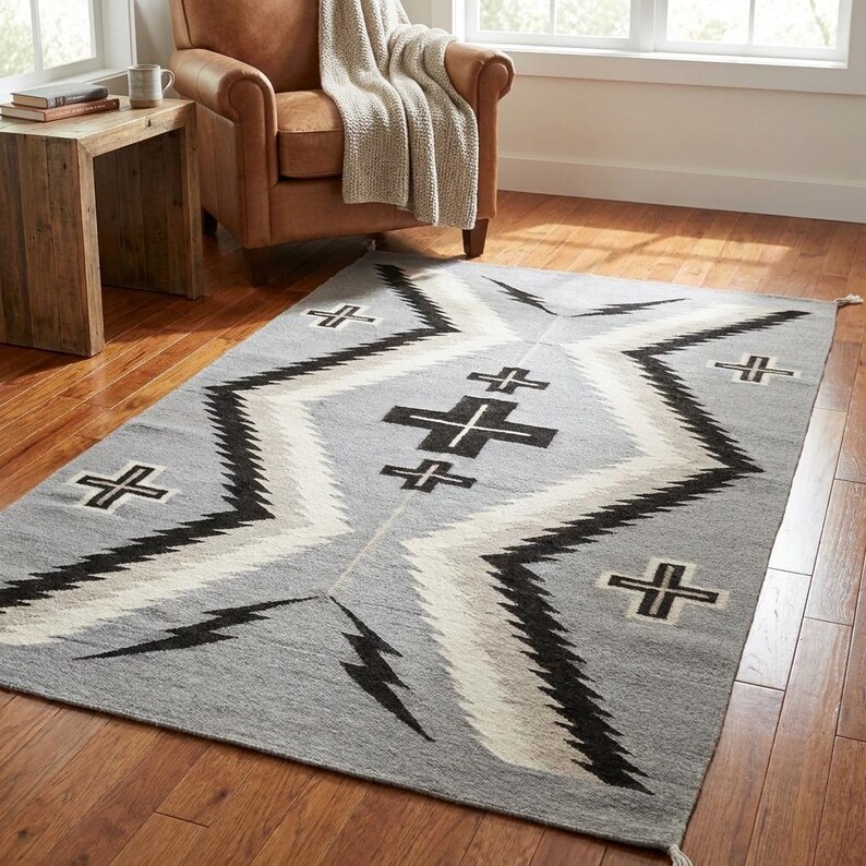 Puede incluir: Una alfombra rectangular gris con un patr&oacute;n geom&eacute;trico. La alfombra presenta l&iacute;neas en zigzag negras, blancas y grises y formas de cruz. La alfombra est&aacute; hecha de un material tejido y tiene flecos en los extremos.