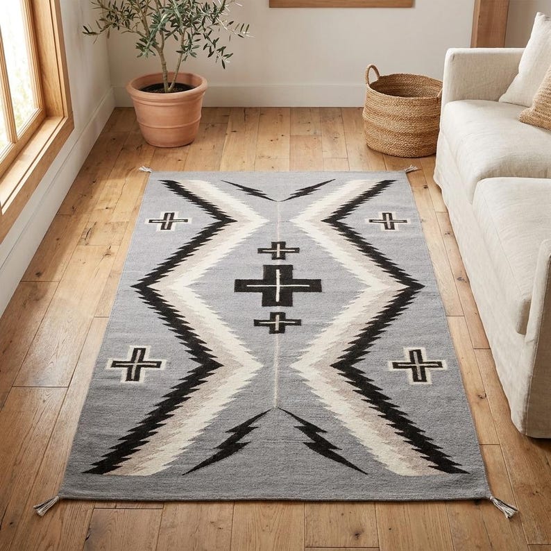 Puede incluir: Una alfombra rectangular gris con un patr&oacute;n geom&eacute;trico. El dise&ntilde;o presenta formas de diamante negras, blancas y beige con motivos de cruz y rel&aacute;mpago. La alfombra tiene borlas en los extremos y est&aacute; colocada sobre un suelo de madera.