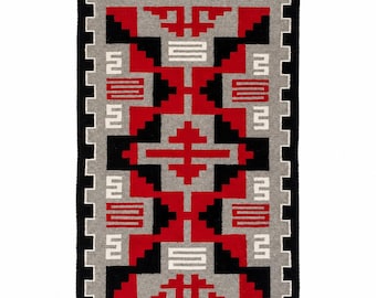 May include: A rectangular woven rug featuring a geometric pattern in red, black, white, and gray. The design includes stepped, zigzag, and angular motifs arranged symmetrically across the surface, finished with small fringe tassels at the corners.