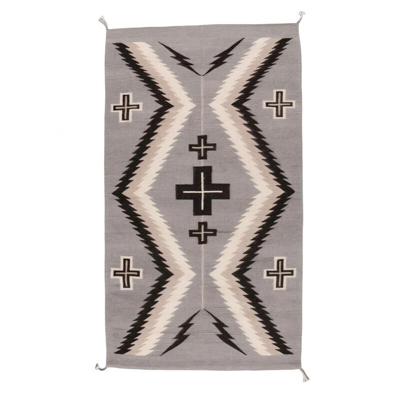 Puede incluir: Alfombra rectangular gris con un dise&ntilde;o geom&eacute;trico. La alfombra presenta una forma de diamante central con zigzags negros, blancos y beige, y cruces negras. La alfombra tiene flecos en los extremos y es adecuada para una sala de estar o dormitorio.