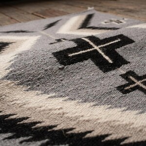 Puede incluir: Primer plano de un textil tejido gris, negro y blanco con un patr&oacute;n geom&eacute;trico. El dise&ntilde;o presenta formas de cruz y l&iacute;neas en zigzag repetidas. El textil tiene una superficie texturizada y parece estar hecho de lana o un material similar.