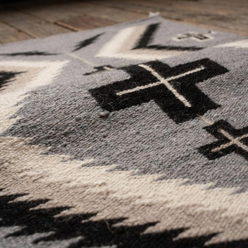 Puede incluir: Primer plano de un textil tejido gris, negro y blanco con un patr&oacute;n geom&eacute;trico. El dise&ntilde;o presenta formas de cruz y l&iacute;neas en zigzag repetidas. El textil tiene una superficie texturizada y parece estar hecho de lana o un material similar.