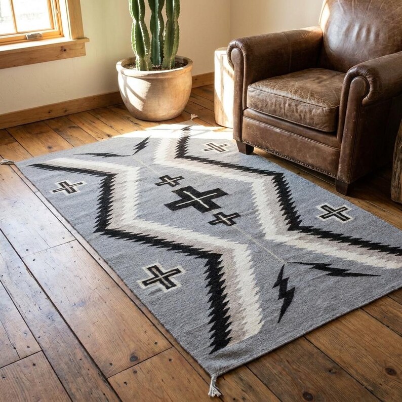 Puede incluir: Una alfombra rectangular gris con un patr&oacute;n geom&eacute;trico. La alfombra presenta un dise&ntilde;o de diamante central con zigzags y cruces negros, blancos y grises. Hay formas de rel&aacute;mpagos en los lados. Hay borlas en los extremos.