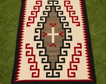May include: A rectangular woven rug featuring a central gray diamond shape with a white cross and red accents. The design includes black and red geometric step patterns on a cream background, framed by a thick black border with small corner tassels.