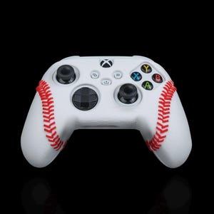 Puede incluir: Mando Xbox blanco con detalles de costuras rojas de béisbol. El mando tiene joysticks negros y botones de colores. El diseño se asemeja a una pelota de béisbol, para los fanáticos del deporte.