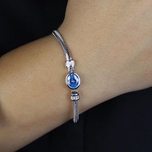 Può includere: Un bracciale d'argento con un ciondolo blu e perline decorative. Il bracciale è su un polso, su sfondo nero. Il ciondolo è ovale, con un centro blu.