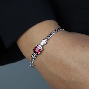 May include: A silver bracelet with a rectangular, deep red gemstone at the center. The bracelet has a snake chain design and is accented with small, clear gemstones. The bracelet is worn on a wrist against a dark background.