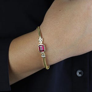 Peut inclure: Bracelet doré orné d'une pierre précieuse rouge foncé de forme rectangulaire. Le bracelet présente un motif de chaîne serpentée texturée avec de petits accents de pierres claires de chaque côté de la pierre. Un bijou classique.