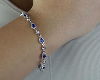 Puede incluir: Una pulsera de plata con piedras preciosas azules en forma de pera y redondas alternas. La pulsera está engastada con pequeñas piedras de acento transparentes. La joya se lleva en la muñeca, sobre un fondo neutro.