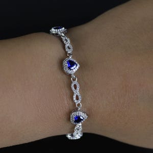 Könnte beinhalten: Ein silbernes Armband mit herzförmigen blauen Edelsteinen und kleinen, klaren Steinen. Das Armband befindet sich an einem Handgelenk vor schwarzem Hintergrund. Das Design umfasst Unendlichkeitszeichen.