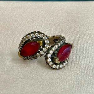 Puede incluir: Dos anillos de estilo antiguo con un centro de piedra roja y un halo de pequeñas piedras transparentes. Los anillos tienen un engaste de color dorado y se exhiben sobre una superficie blanca.
