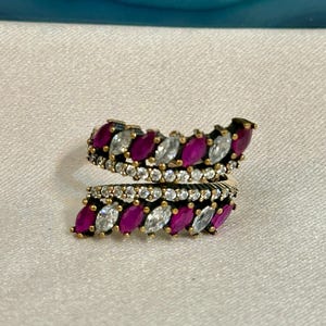 May include: A silver-toned ring with a wrap-around design. The ring is adorned with alternating rows of oval-cut, deep pink and clear gemstones. The gemstones are set in a detailed, textured band.