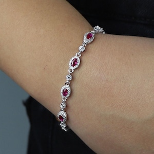Puede incluir: Una pulsera plateada con piedras preciosas rojas ovaladas y piedras claras redondas más pequeñas. La pulsera se luce en una muñeca, mostrando el diseño detallado y los detalles brillantes. La joya se presenta sobre un fondo oscuro.