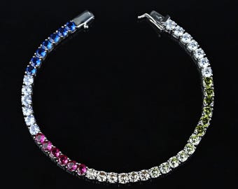 Può includere: Un braccialetto tennis con un gradiente di pietre preziose colorate. Il braccialetto passa dal blu al bianco, poi al rosa e infine al verde. Le pietre preziose sono incastonate in un metallo color argento.