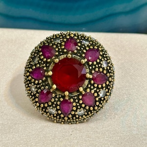 Puede incluir: Un anillo redondo de estilo antiguo con una gran gema roja central rodeada de piedras rojas más pequeñas en forma de lágrima. El engaste del anillo es dorado con pequeñas piedras de acento transparentes y una banda de metal oscuro texturizado.
