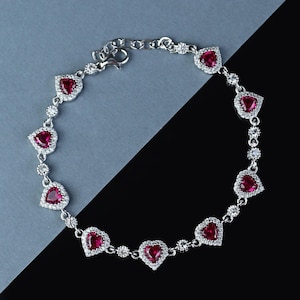 Puede incluir: Una pulsera de plata con piedras preciosas rojas en forma de corazón, cada una rodeada de pequeños cristales transparentes. La pulsera tiene una cadena delicada y un cierre seguro. El diseño es elegante y adecuado.