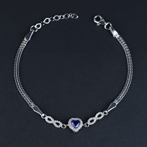 Könnte beinhalten: Ein silbernes Armband mit einem herzförmigen blauen Edelstein. Das Armband hat ein filigranes Kettendesign mit Unendlichkeitszeichen-Akzenten und einen Verschluss. Der Schmuck wird vor einem dunklen Hintergrund präsentiert.