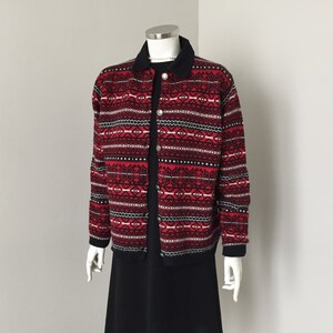 May include: A red, black, and white patterned cardigan with a black collar and silver buttons. The cardigan is paired with a black dress. The outfit is displayed on a mannequin.
