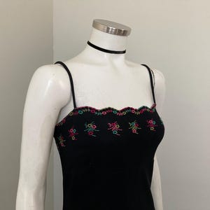 May include: A black sleeveless dress featuring thin spaghetti straps and a scalloped neckline. The upper bodice is decorated with a horizontal row of embroidered floral motifs in vibrant shades of pink, teal, and green against the black fabric.
