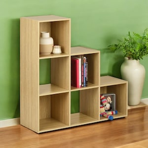 May include: A light wood 6-cube shelving unit with a staggered design. The shelf unit has a vase with a decorative object on the top shelf, books on the middle shelf, and a stuffed toy on the bottom shelf.