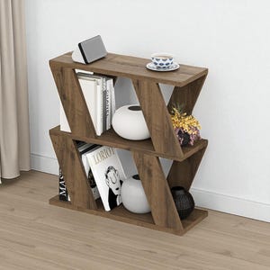 May include: A brown wooden bookshelf with a distinctive zigzag design. The shelves display books, white vases, a small speaker, a teacup, and a decorative plant. The bookshelf is positioned against a white wall and a light brown floor.