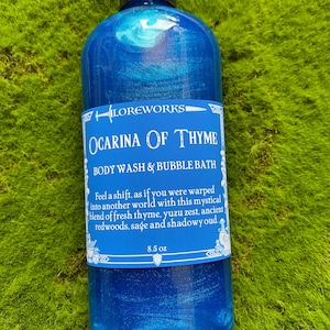 May include: A blue bottle of Ocarina of Thyme body wash and bubble bath. The bottle is 8.5 oz and has a label with a white background and black text. The text on the label reads "LOREWORKS OCARINA OF THYME BODY WASH & BUBBLE BATH Feel a shift, as if you were warped into another world with this mystical blend of fresh thyme, yuzu zest, ancient redwoods, sage and shadowy oud. 8.5 oz"