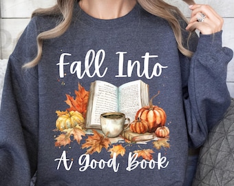Op de afbeelding: Een marineblauw sweatshirt met de tekst "Fall Into A Good Book" in wit. Het ontwerp bevat een aquarel-illustratie van een open boek, een kopje koffie en pompoenen met herfstbladeren.