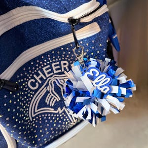 May include: A blue glittery backpack with white trim and the word CHEER printed above a stingray graphic. Attached to the zipper is a blue and white pom-pom keychain featuring a blue acrylic name tag with the name Ella in white script.