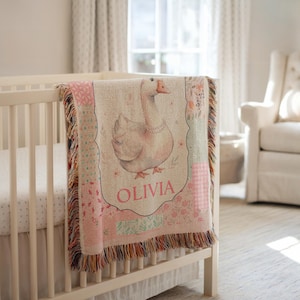 Può includere: Una coperta per bambini con un'illustrazione di un'oca e il nome "OLIVIA" in rosa, drappeggiata su una culla. La coperta presenta un design patchwork con motivi floreali e geometrici e un bordo con frange.