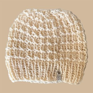 May include: Cream-colored knitted beanie hat with a textured pattern. The hat has a ribbed brim and a small metal tag that reads "THE 6TH STITCH". The hat is made of a soft, warm material.