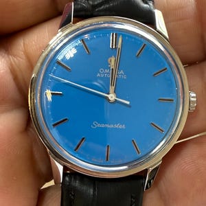 Könnte beinhalten: Eine silberne Omega Seamaster Automatic Armbanduhr mit blauem Zifferblatt und schwarzem Lederarmband.