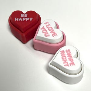 May include: Three heart-shaped trinket boxes in red, pink, and white. The red box says "BE HAPPY", the pink box says "I LOVE YOU", and the white box says "SWIPE RIGHT" in pink lettering. The boxes are on a white surface.