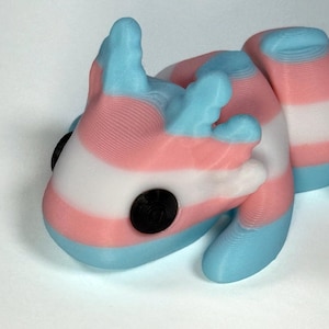 Può includere: Statuetta stampata in 3D di una lucertola stilizzata, con una combinazione di colori a strisce blu chiaro, rosa e bianco. La lucertola ha due occhi neri circolari e una superficie strutturata.