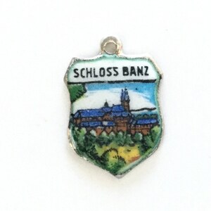May include: A silver charm in the shape of a shield with a painted image of a castle and the text "SCHLOSS BANZ".
