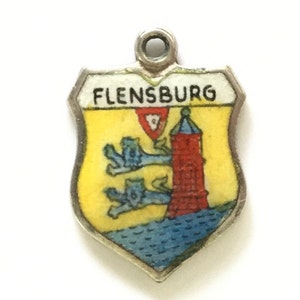 Puede incluir: Un amuleto de plata en forma de escudo con fondo amarillo y una torre roja. El amuleto tiene detalles azules y verdes y el texto "FLENSBURG" en la parte superior.