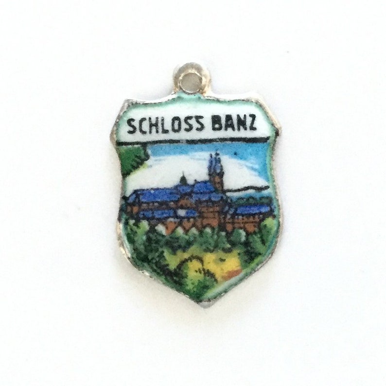May include: A silver charm in the shape of a shield with a painted image of a castle and the text "SCHLOSS BANZ".