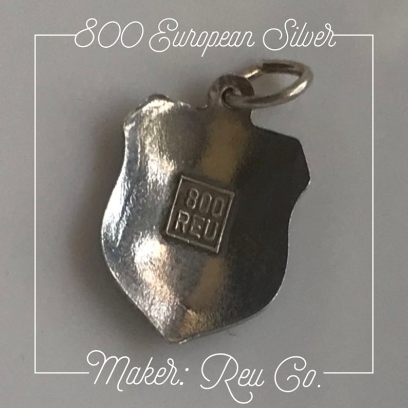 May include: Silver charm in the shape of a shield with the text "800 REU" on the back. The charm is marked "800 European Silver" and "Maker:  Reu Co."