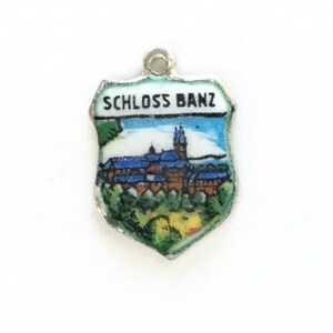 May include: A silver charm in the shape of a shield with a blue and white image of a castle and the text "SCHLOSS BANZ" on the top.