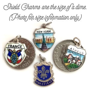 May include: Four enamel shield charms with different designs. The charms are silver-toned and feature images of the Statue of Liberty, the Acropolis in Athens, the French flag, and a fleur-de-lis. The text "NEW YORK", "ATHENS ACROPOLIS", "FRANCE", and "WAKEFIELD" is visible on the charms. The charms are approximately the size of a dime.