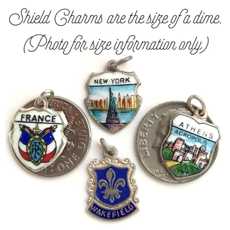 May include: Four enamel shield charms with different designs. The charms are silver-toned and feature images of the Statue of Liberty, the Acropolis in Athens, the French flag, and a fleur-de-lis. The text "NEW YORK", "ATHENS ACROPOLIS", "FRANCE", and "WAKEFIELD" is visible on the charms. The charms are approximately the size of a dime.