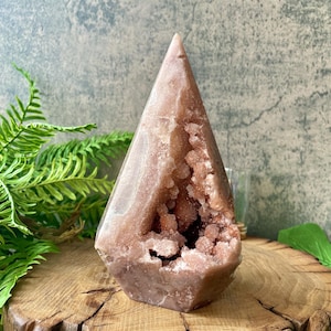 May include: A pink crystal geode with a pointed top and a hollowed-out center filled with smaller crystals. The geode sits on a wooden surface, with green ferns and leaves in the background. The crystal is approximately 6 inches tall.