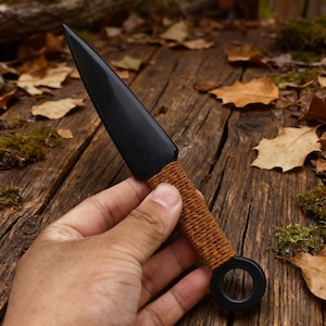 May include: A black kunai knife with a sharp, pointed blade. The handle is wrapped in brown twine, and a black ring is at the base. The knife is displayed on a weathered wooden surface with fallen leaves.