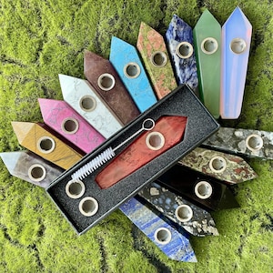 May include: A collection of gemstone pipes in various colors, including pink, blue, green, and brown, arranged on a bed of green moss. Each pipe has a metal bowl and a pointed end. A red pipe and cleaning brush are in a black box.